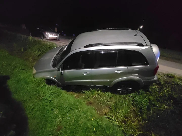 Srebrny SUV zjechał z drogi i stoi w przydrożnym rowie nocą, w tle widać inny samochód na jezdni.