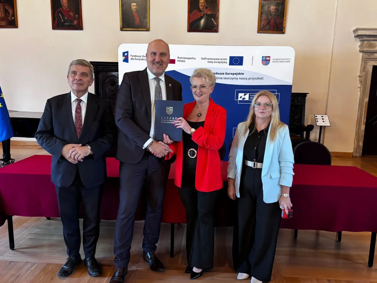 Cztery osoby pozują podczas uroczystości wręczenia dokumentu w sali z banerem Funduszy Europejskich dla Świętokrzyskiego.