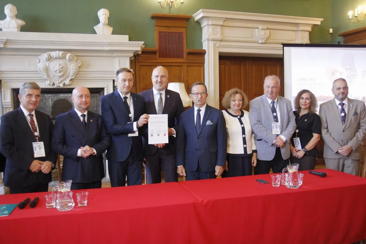 Grupa osób w formalnym stroju stoi za czerwonym stołem w reprezentacyjnej sali, dwaj mężczyźni trzymają podpisany dokument.