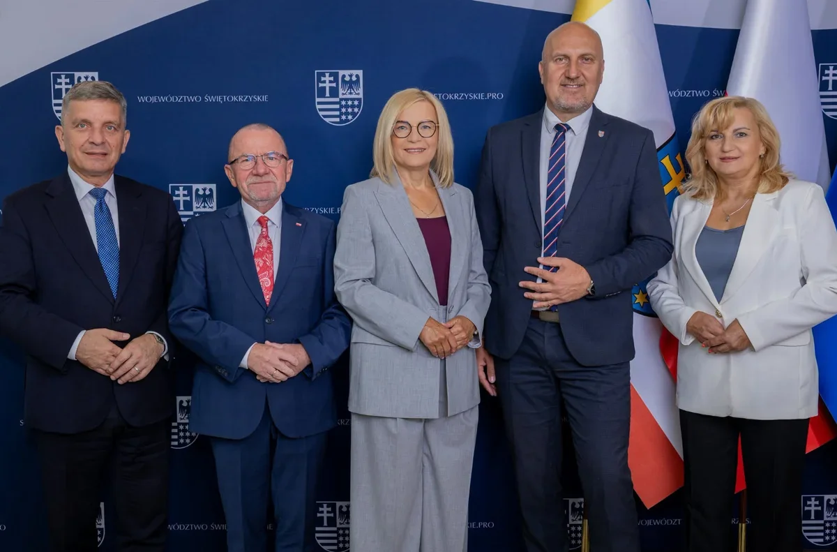 Pięcioro osób w eleganckim stroju biznesowym pozuje do grupowego zdjęcia przed ścianką z herbem i napisami województwa świętokrzyskiego oraz polskimi flagami.