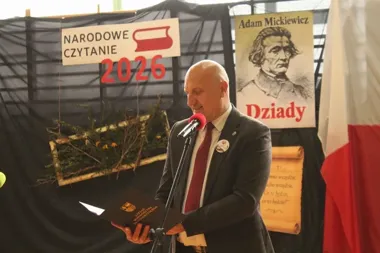 Mężczyzna w garniturze czyta z książki przy mikrofonie podczas Narodowego Czytania 2026, w tle plakat Adama Mickiewicza z napisem „Dziady”.