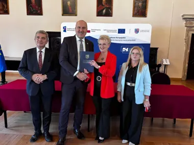 Cztery osoby pozują podczas uroczystości wręczenia dokumentu w sali z banerem Funduszy Europejskich dla Świętokrzyskiego.