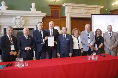 Grupa osób w formalnym stroju stoi za czerwonym stołem w reprezentacyjnej sali, dwaj mężczyźni trzymają podpisany dokument.