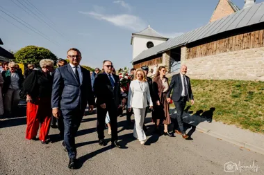Grupa osób w eleganckich strojach przemierza ulicę przed drewnianym kościołem w słoneczny dzień.